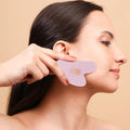 Woman using pink facial gua sha tool for skincare routine, promoting glowing healthy complexion